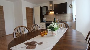Une table à manger agrémentée d’un chemin de table à motifs floraux, entourée de chaises en bois. On aperçoit à l’arrière-plan une cuisine avec des meubles sombres et un dosseret carrelé.