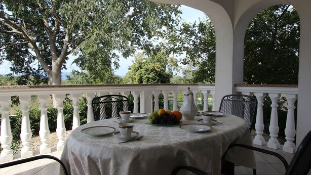 Une table ronde dressée pour un repas, recouverte d’une nappe blanche, entourée de balustrades blanches, et offrant une vue sur des arbres et des montagnes en arrière-plan.