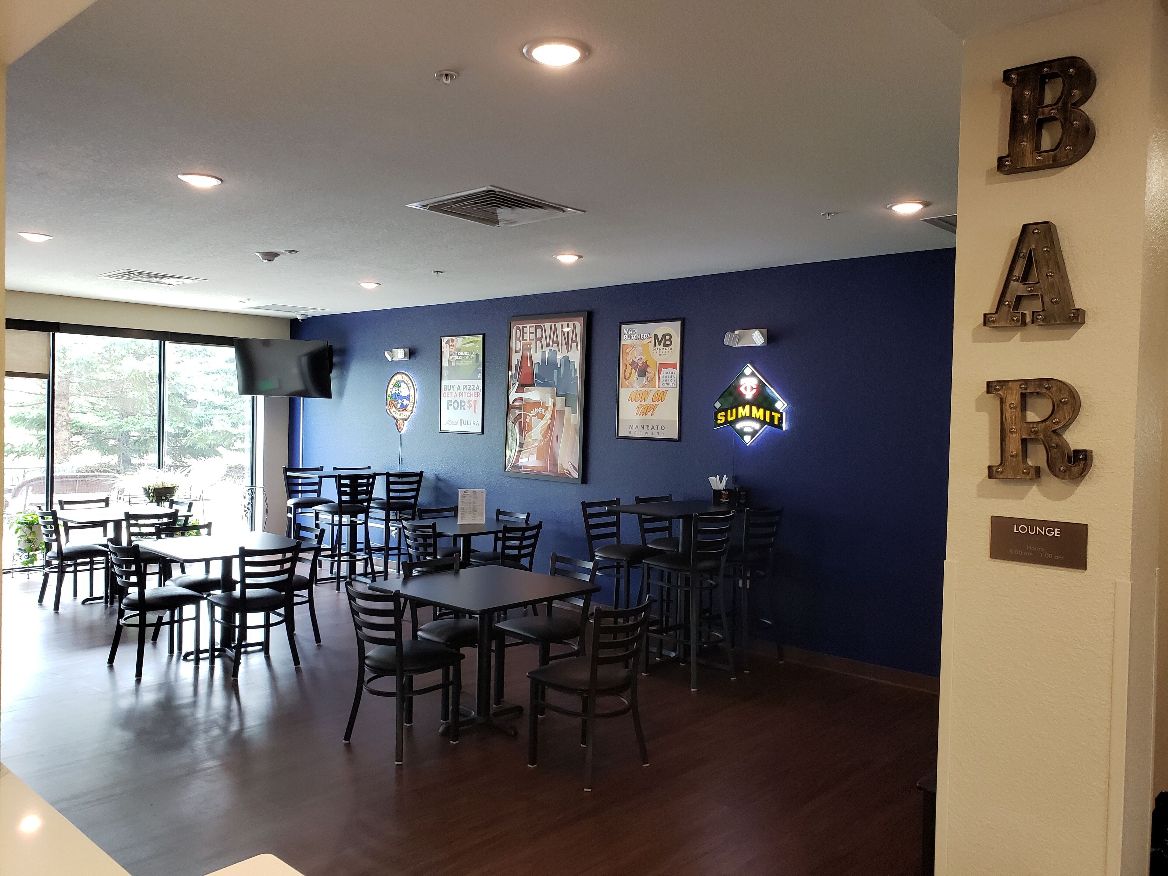 A restaurant interior with multiple tables and chairs, a bar area with illuminated letters, and a large window providing natural light.