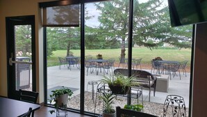 A room with a large sliding glass door leading to an outdoor patio with tables and chairs, potted plants, and a view of trees and grass.
