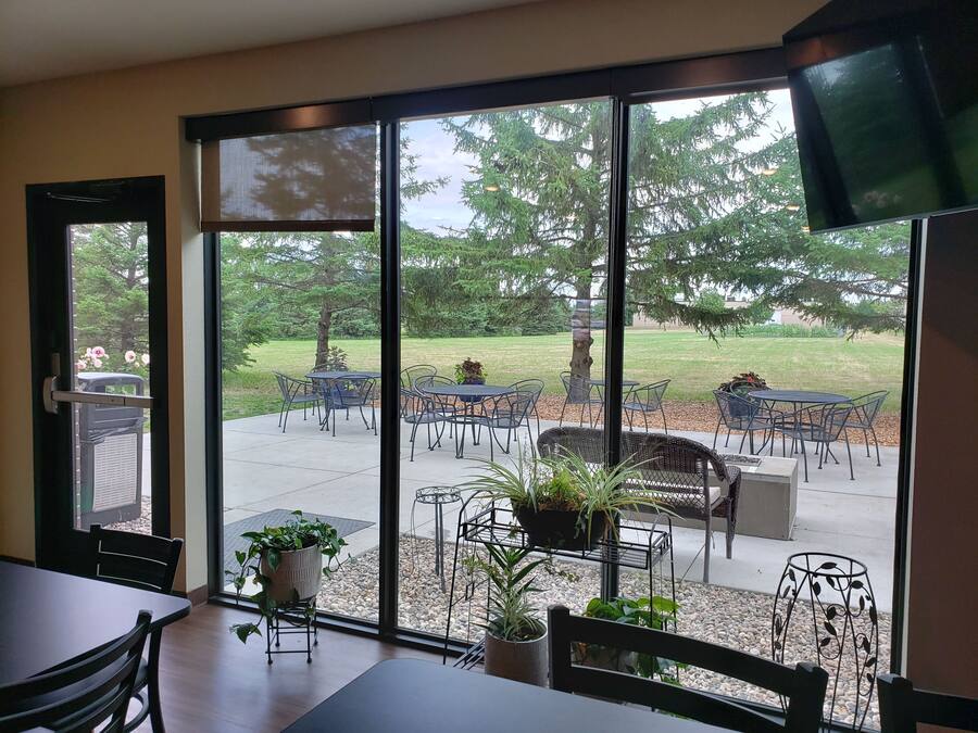 Una habitación con un gran ventanal corredero que da a un patio exterior con mesas y sillas, plantas en macetas y vistas a árboles y césped.