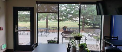 A modern indoor dining area with a large sliding glass door leading to an outdoor patio. There are several tables and chairs arranged for dining, and a flat-screen TV mounted on the wall.