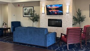A lobby with a blue sofa, red chairs, a TV mounted on a textured wall, and a fireplace below it.