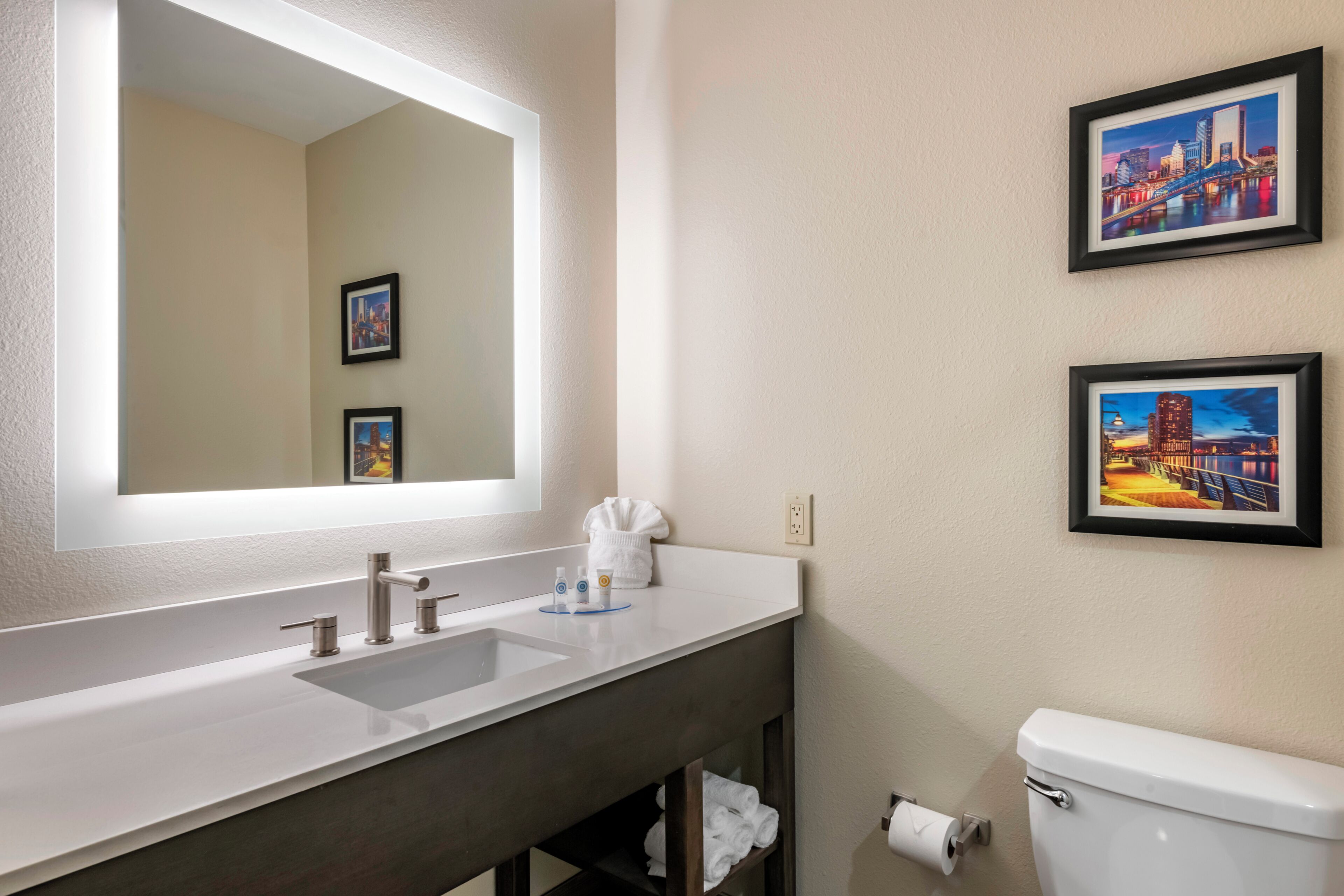 Un baño con un espejo grande, dos lavamanos, un inodoro y dos cuadros en la pared.