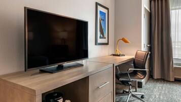 A hotel room with a flat-screen TV on a wooden desk, a chair, a desk lamp, and a framed picture on the wall.