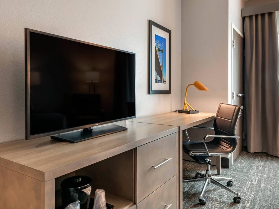 Ett hotellrum med en platt-TV på ett skrivbord i trä, en stol, en skrivbordslampa och en inramad tavla på väggen.