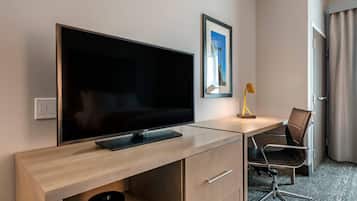 A hotel room with a large flat-screen TV on a wooden desk, a framed picture on the wall, a desk lamp, a black office chair, and a door with curtains.