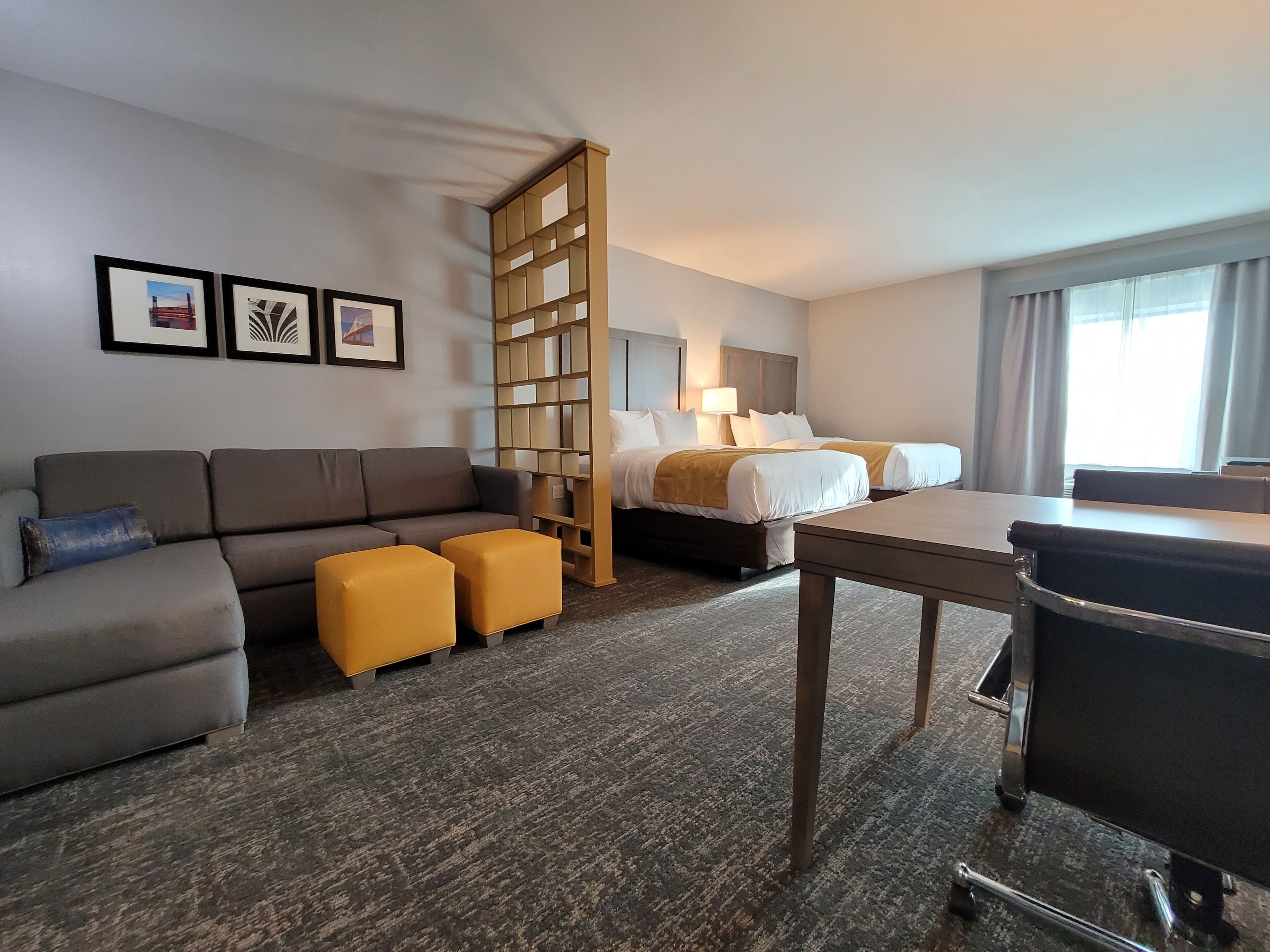 A hotel room with a sofa, two yellow ottomans, a bed with white linens, a desk, and a bookshelf.