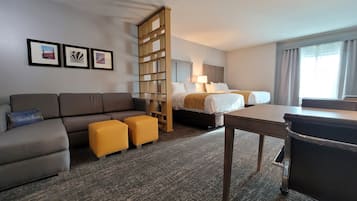 Una habitación de hotel con un sofá, dos otomanos amarillos, una cama con ropa blanca, un escritorio y una estantería.