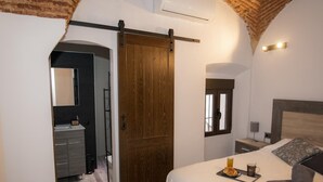 Une chambre avec une porte en bois, un lit avec une tête de lit, une table de chevet, une commode, une fenêtre et une salle de bain visible à travers une porte ouverte.