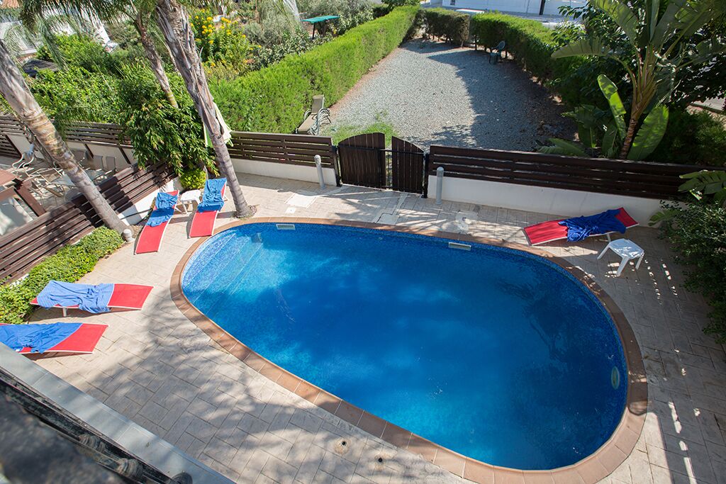 Une piscine entourée d’une clôture en bois et de verdure, avec des chaises longues rouges et bleues.