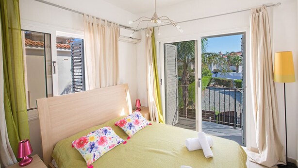 Una camera d'albergo con un letto, un comodino, una lampada, un balcone con vista e una finestra con tende.