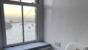 A room with a view of the sea through a window, a bed with white bedding, and a cushion with anchor patterns.