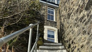 A stone staircase leading up to a building with a window, surrounded by vegetation and a rocky wall.