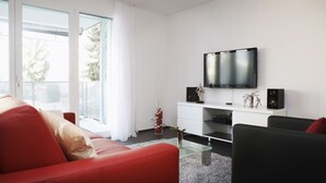 Ein modernes Wohnzimmer mit einem roten Sofa, einem schwarzen Sessel und einem Glastisch. Ein Flachbildfernseher ist an der Wand über einer weißen Unterhaltungsanlage mit Lautsprechern angebracht.