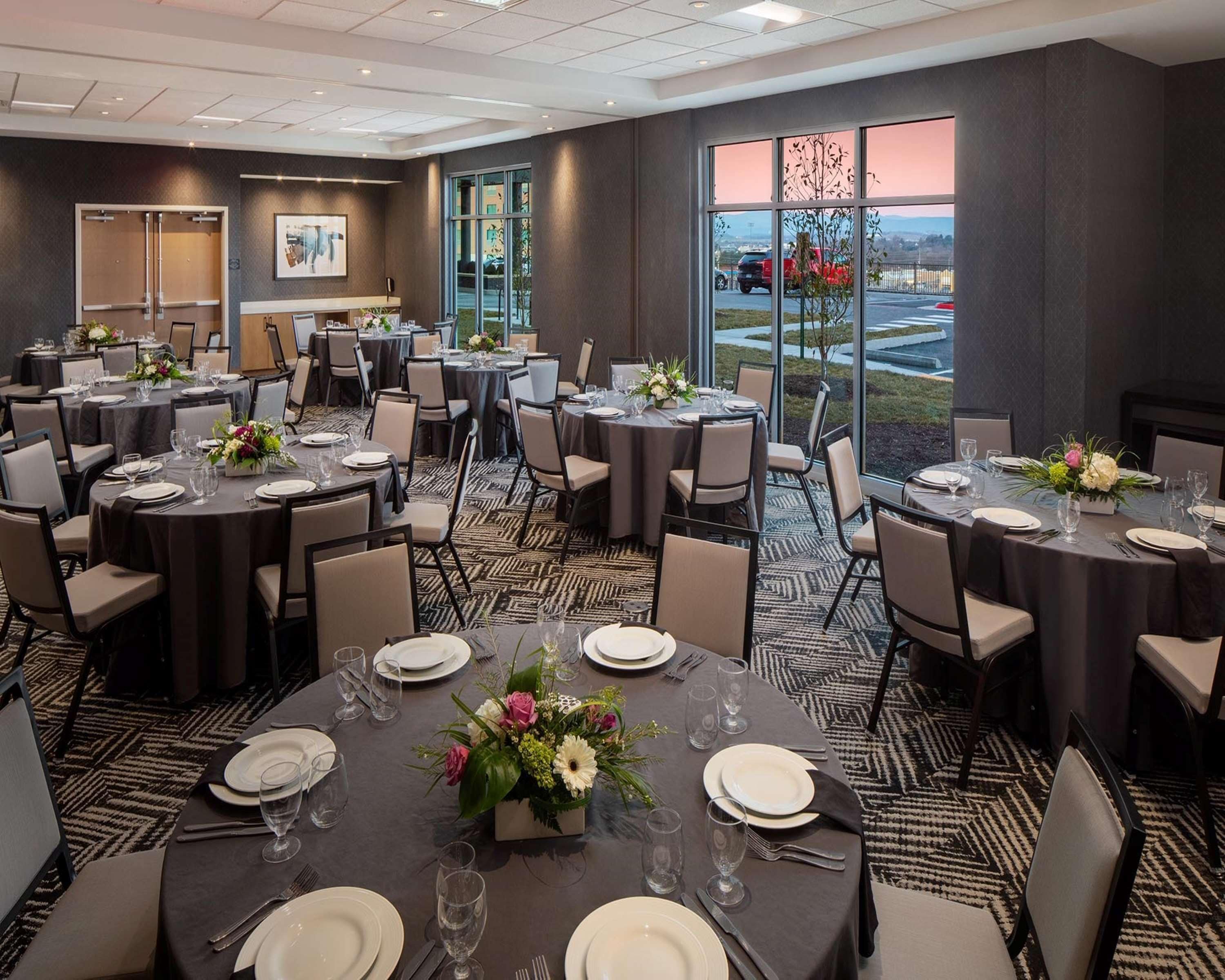 Une salle de conférence équipée de plusieurs tables dressées pour un repas, de grandes fenêtres donnant sur le parking et d’un sol recouvert de moquette.