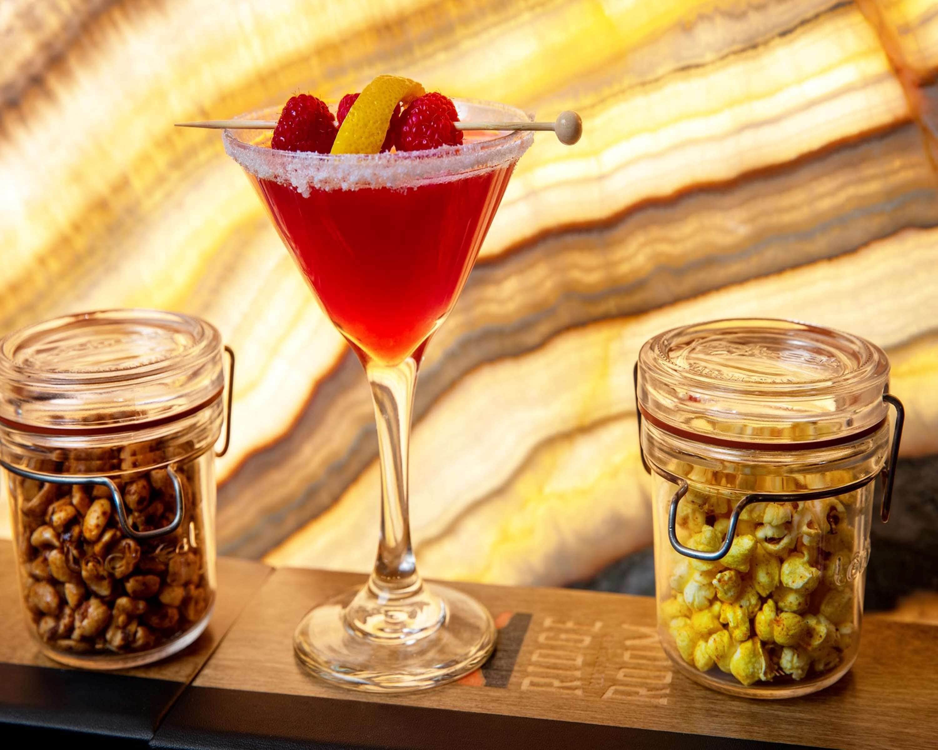 A martini glass with a red cocktail, garnished with raspberries and an orange peel, placed on a wooden surface with two glass jars containing popcorn and nuts.
