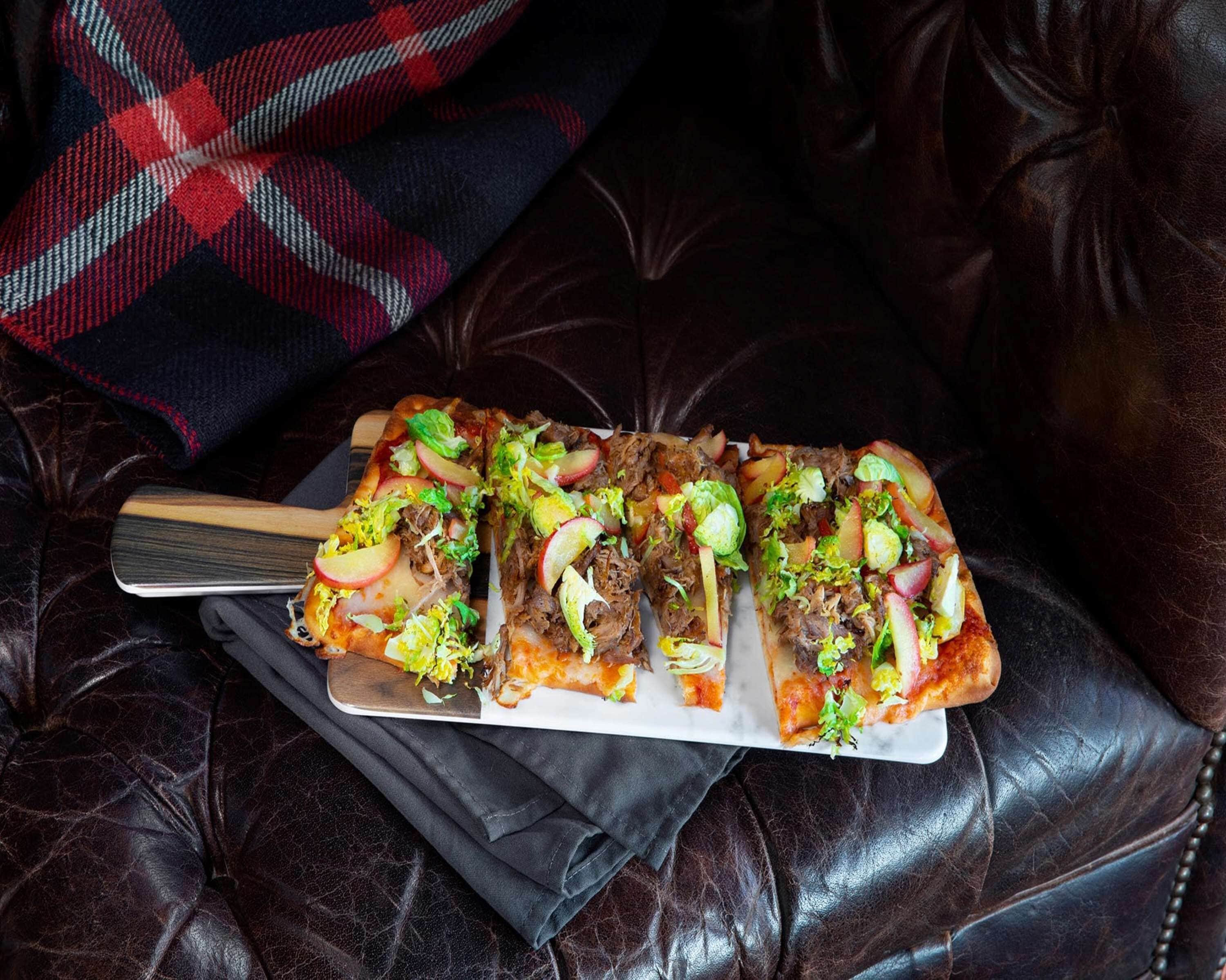 En rektangulær pizza med forskellige toppings på en hvid tallerken, placeret på en mørk lædersofa med et ternet tæppe i baggrunden.