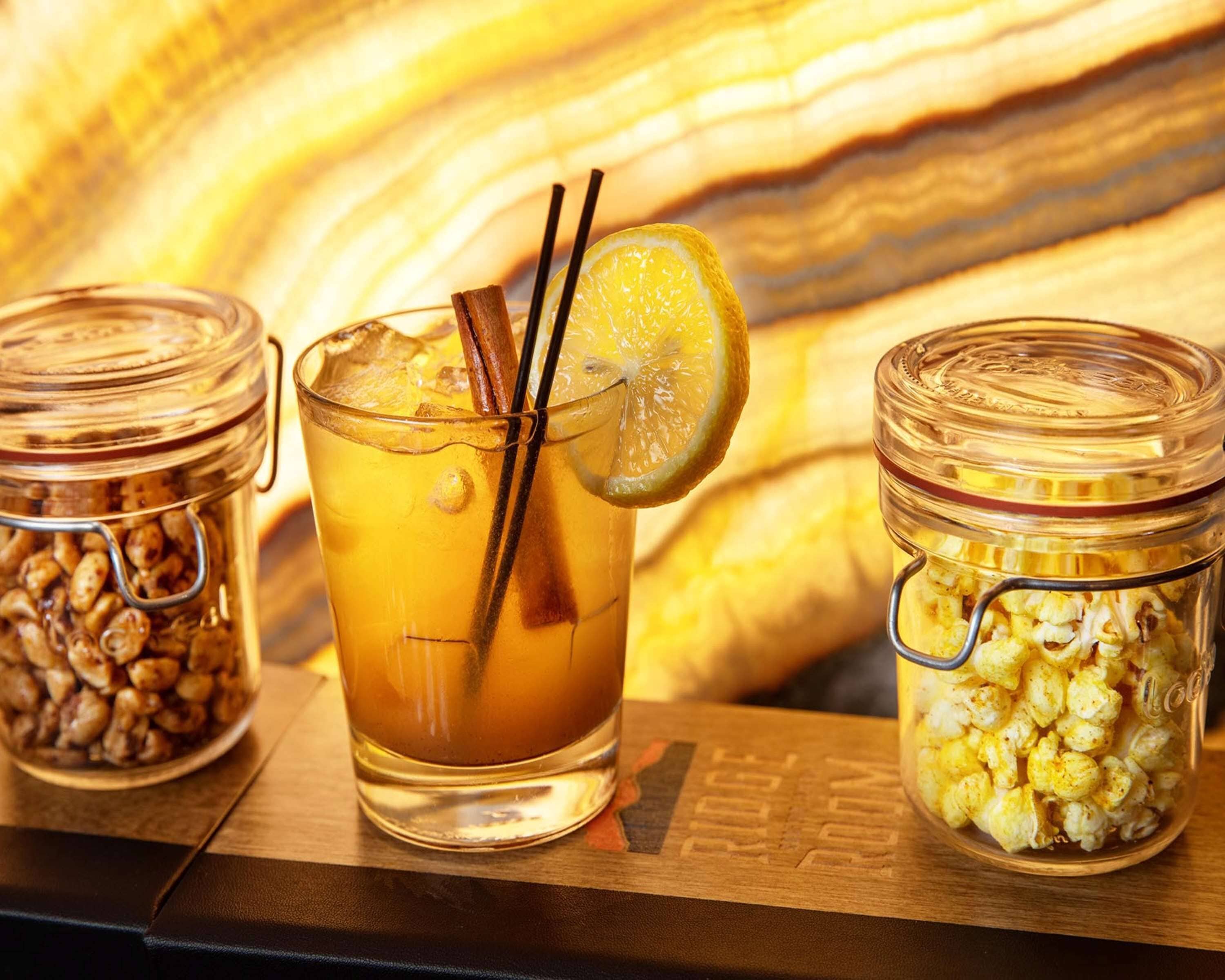 A glass of amber-colored beverage with a lemon slice and cinnamon sticks, accompanied by two jars of popcorn and a jar of nuts.