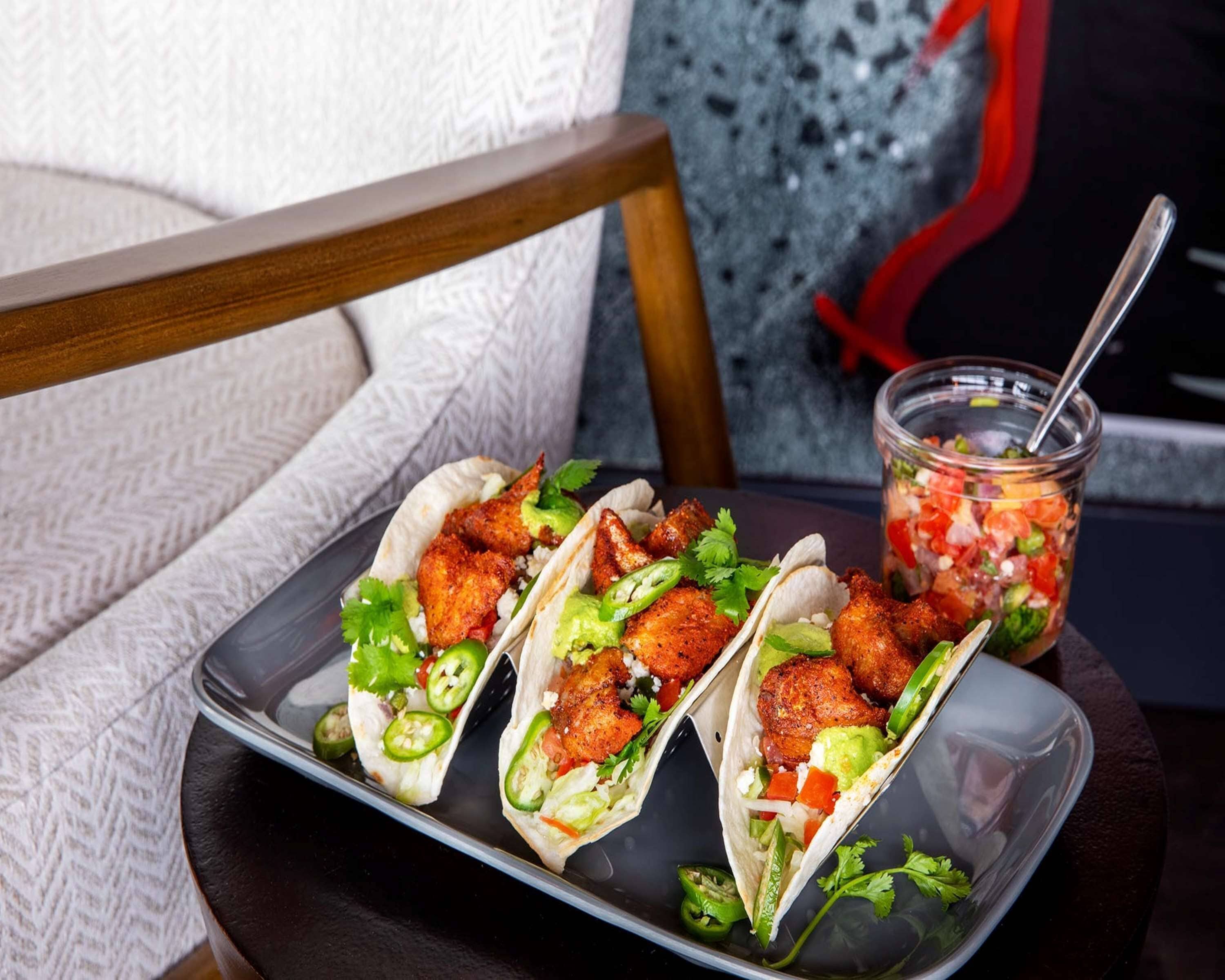 Three tacos on a tray with a glass of salsa on a table.
