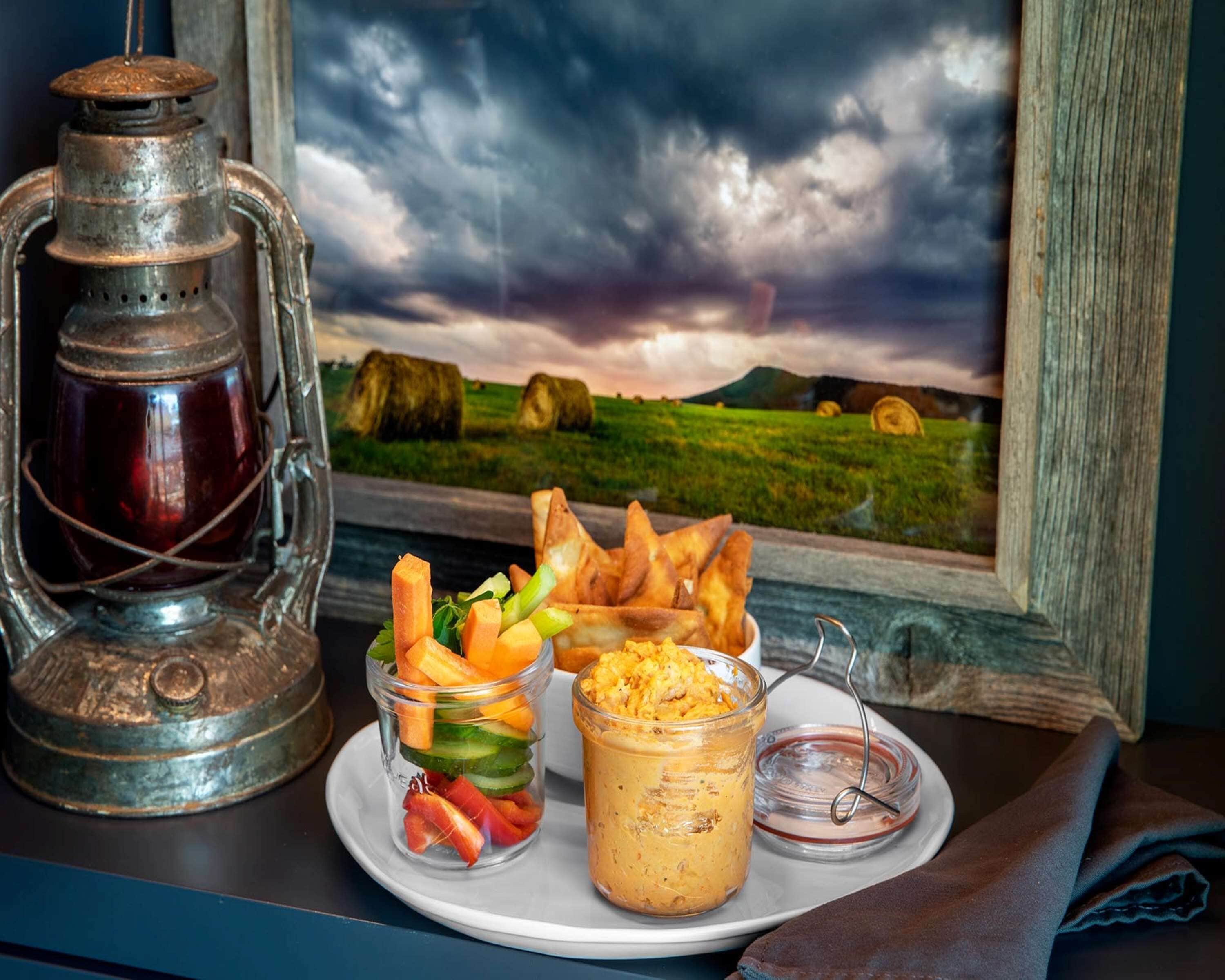 Una mesa rústica con un farol, un plato de comida que incluye verduras y pan, y un frasco de salsa, todo frente a una vista panorámica del campo.