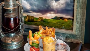 A rustic table setting with a lantern, a plate of food including vegetables and bread, and a jar of dip, set against a scenic countryside view.