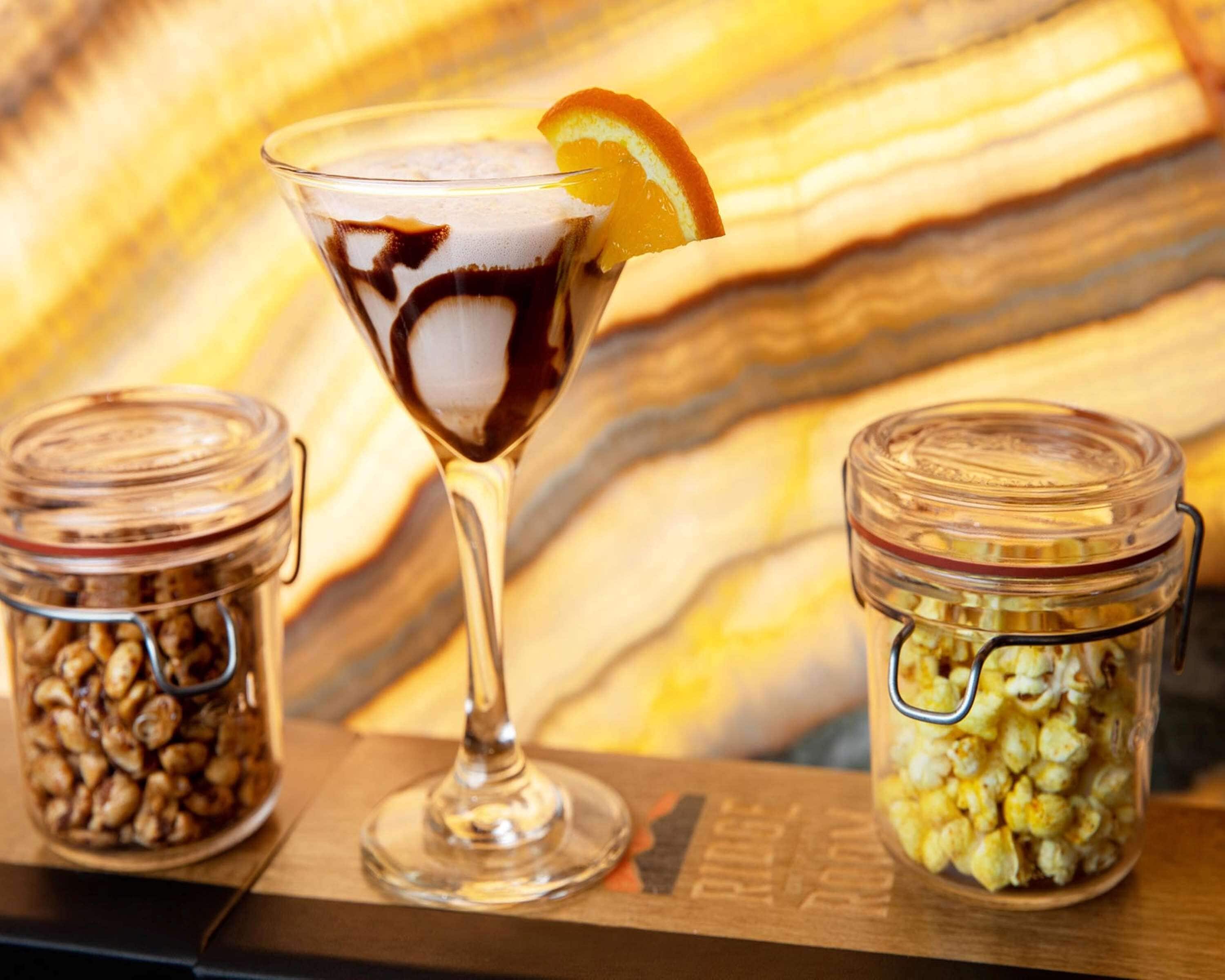 A martini glass with a chocolate syrup swirl and an orange garnish, two glass jars with popcorn and nuts, and a wooden surface with a striped pattern.
