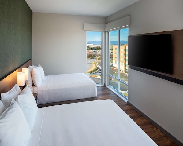 Habitación de hotel con dos camas, un ventanal grande con vistas a la ciudad y un televisor montado en la pared.
