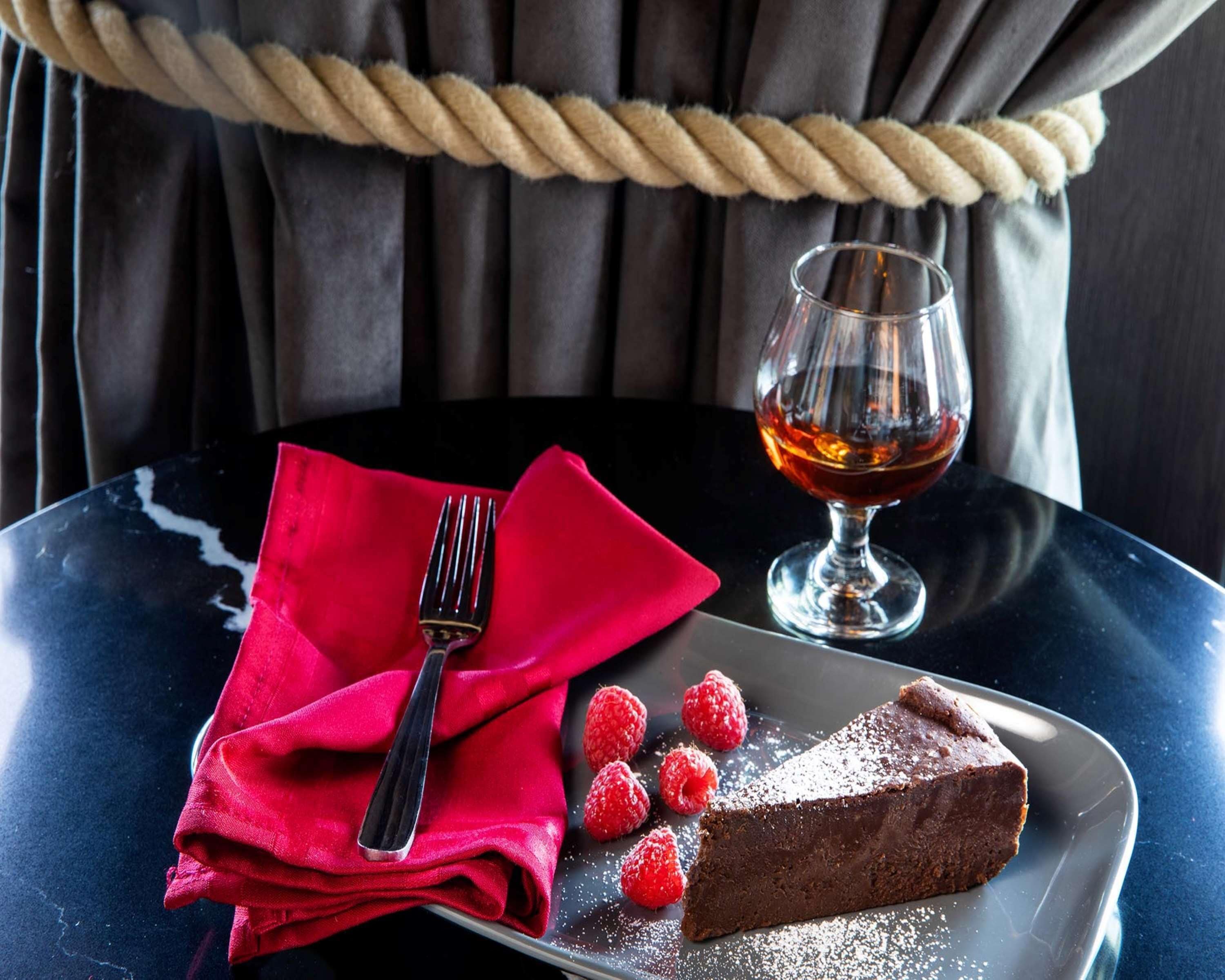 Un plato de postre con pastel de chocolate, frambuesas y un vaso de líquido ámbar sobre una mesa.