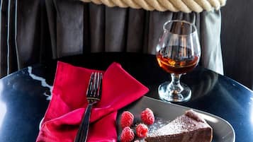 Une assiette à dessert avec un morceau de gâteau au chocolat, des framboises et un verre d’un liquide ambré, le tout posé sur une table.