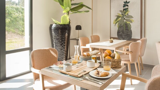 Une table mise pour le petit-déjeuner, avec une nappe, de la vaisselle, des boissons et un vase rempli de plantes vertes.