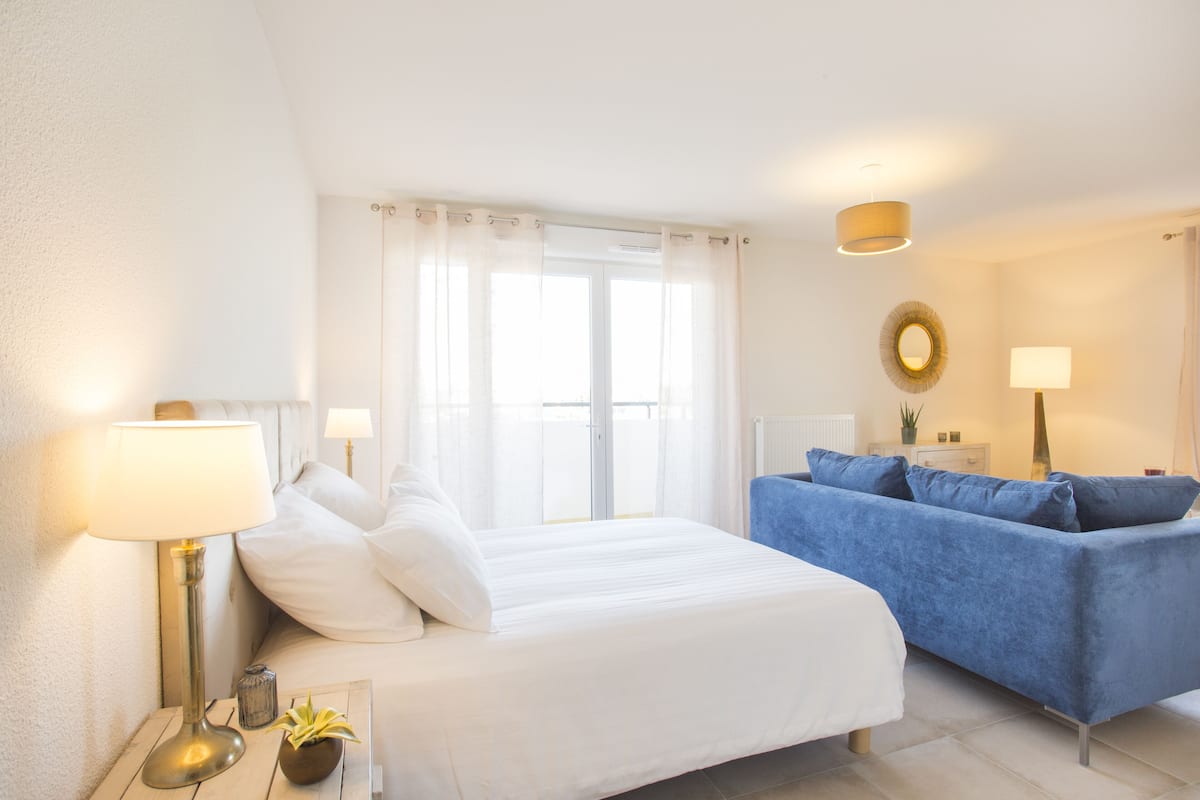 A bedroom with a bed, a blue sofa, two bedside lamps, a window with curtains, and a wall-mounted mirror.