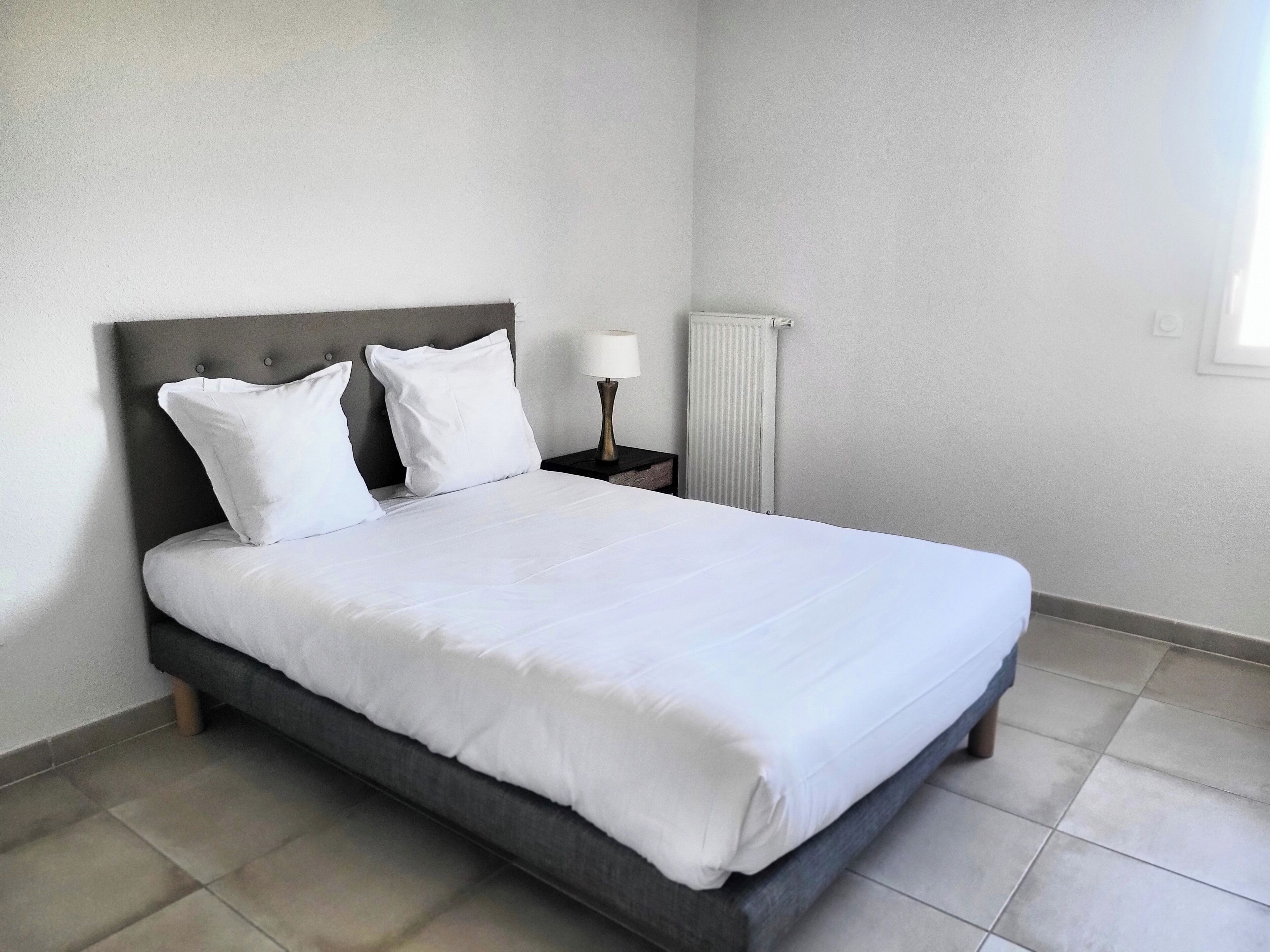 A neatly made bed with white linens in a room with a grey headboard, a bedside table with a lamp, and a radiator.
