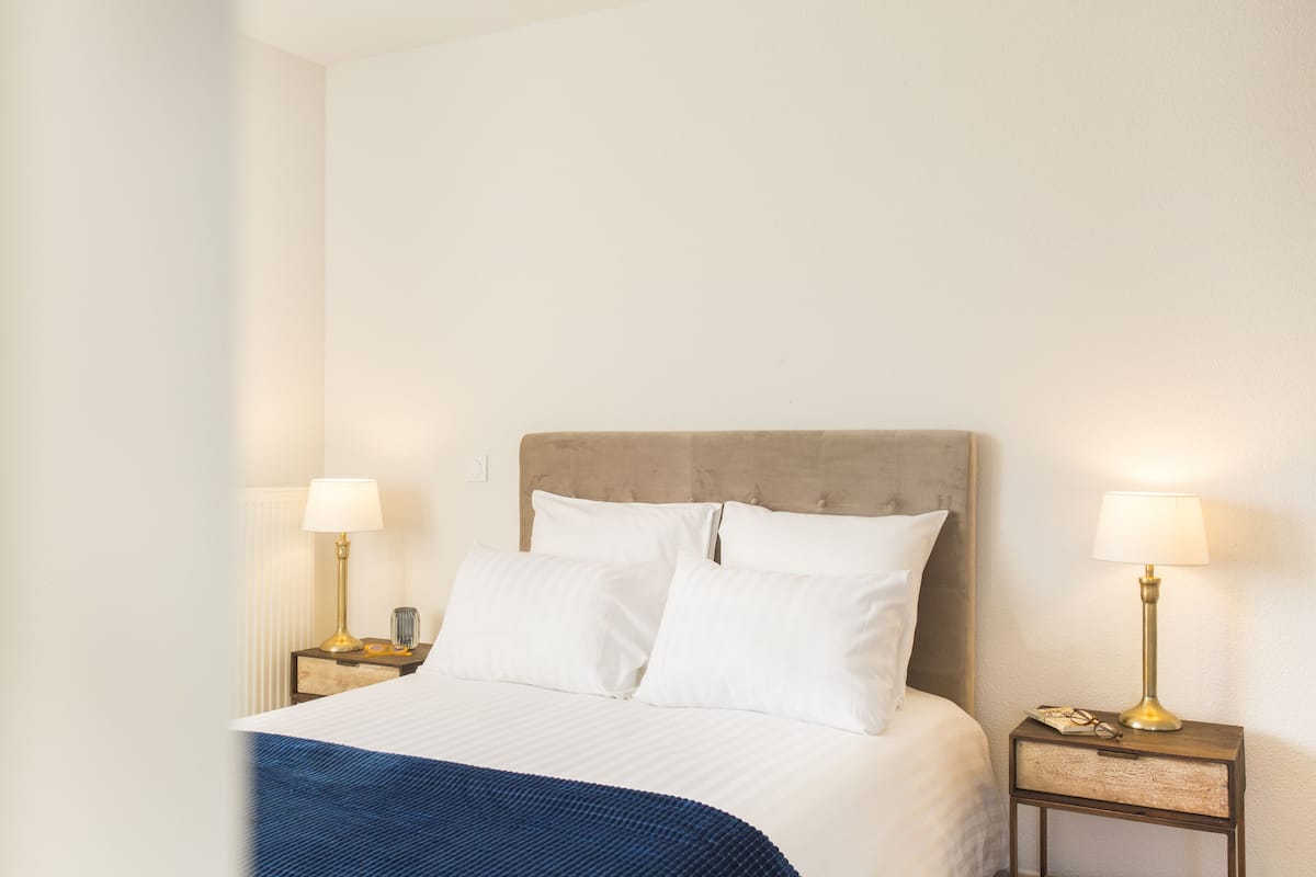 A neatly made bed with white linens and a blue throw blanket, flanked by two bedside tables with lamps and a small decorative item on one.