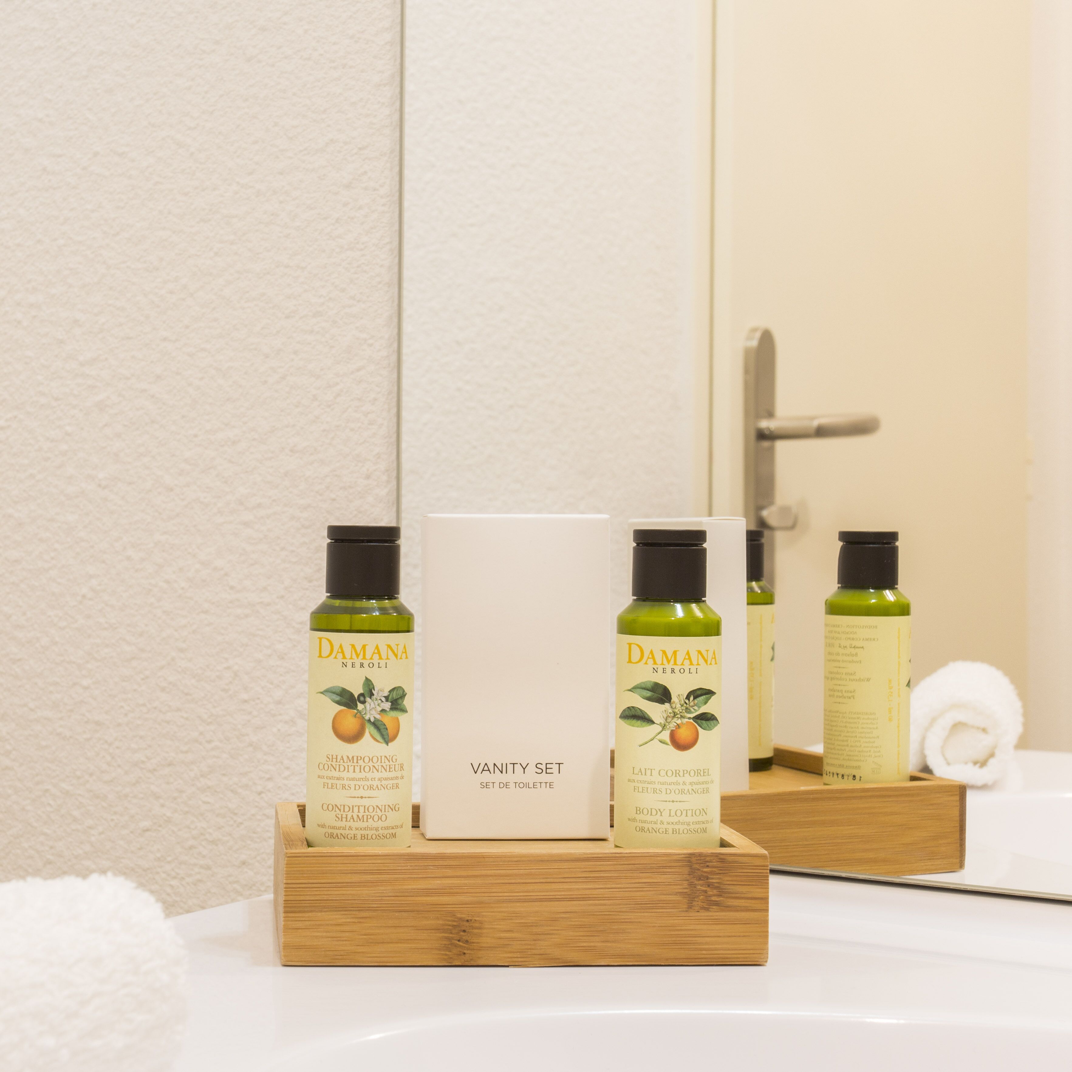 Un plateau en bois contenant trois flacons de shampoing et de lotion pour le corps à la fleur d’oranger Damana, ainsi qu’une trousse de toilette avec un miroir et une serviette.