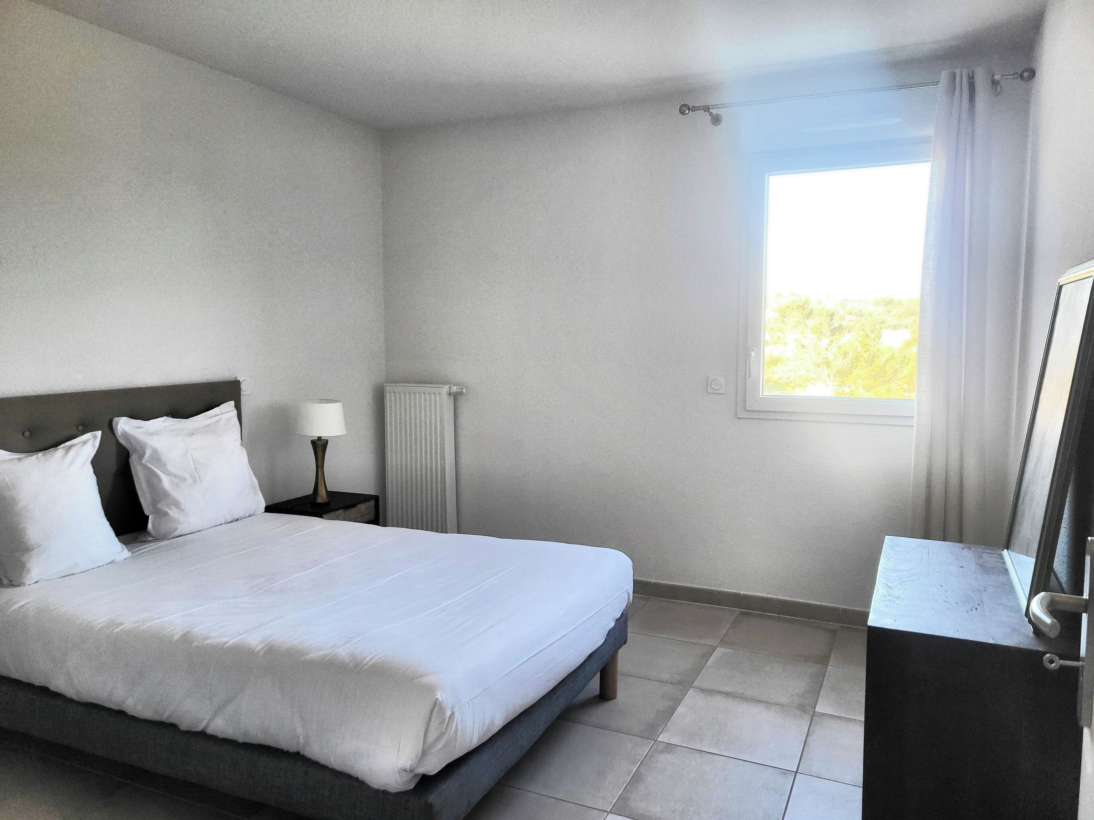 A hotel room with a bed, a nightstand with a lamp, a radiator, and a window with curtains.