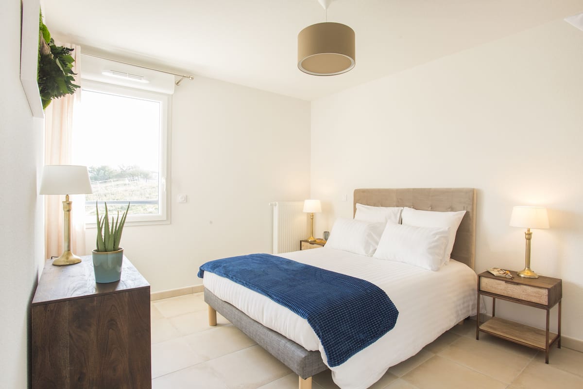 A bedroom with a bed, bedside tables, a nightstand, a potted plant, and a window.