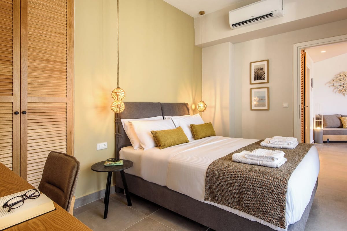 A hotel room with a bed, bedside table, and a chair. There is a wall-mounted air conditioner and a framed picture on the wall.
