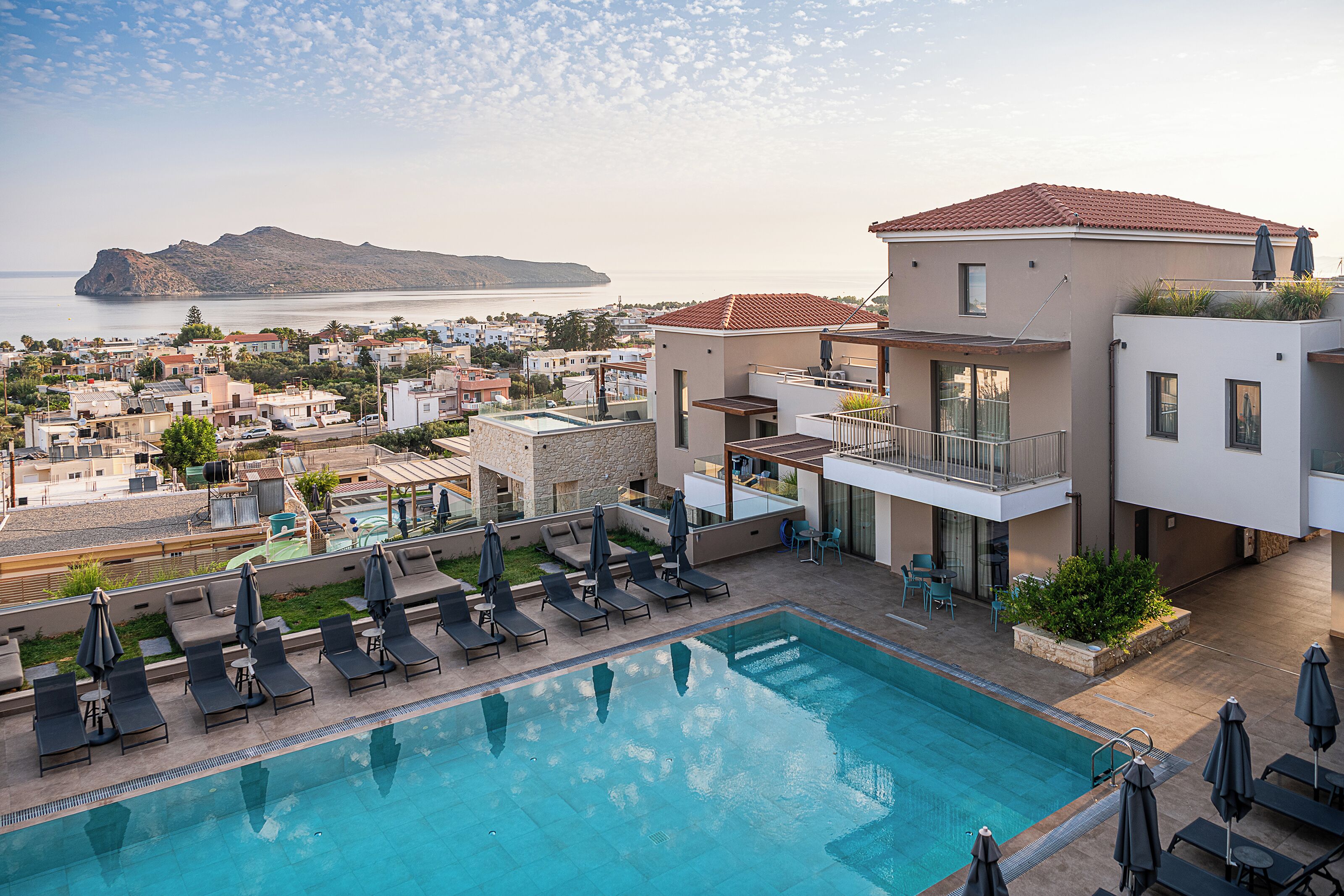 Une piscine avec des chaises longues et des parasols, une vue sur la mer et un bâtiment avec un toit recouvert de tuiles rouges.