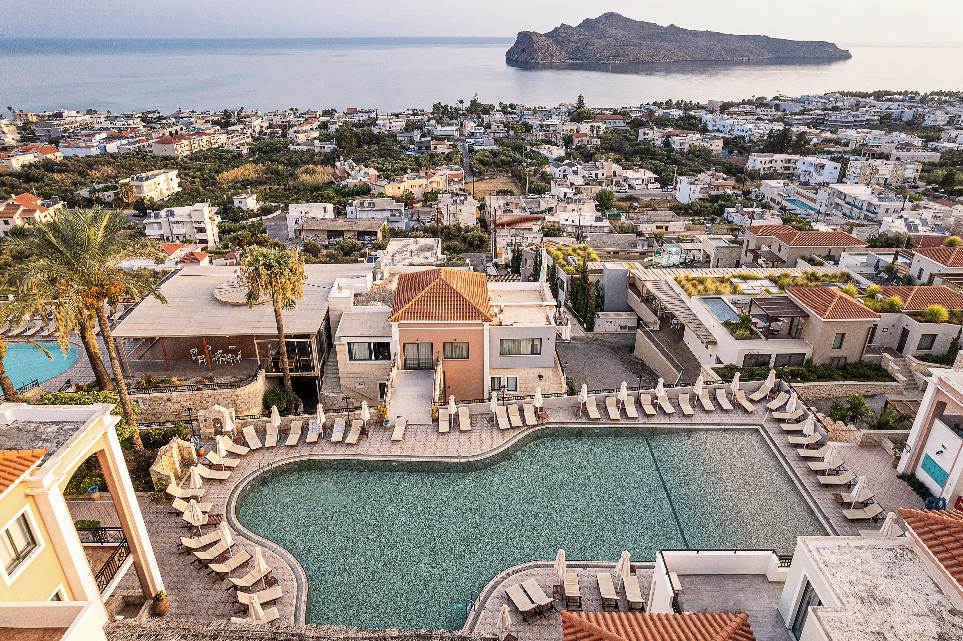 A resort with a large pool surrounded by sun loungers, a view of the sea, and a mountainous backdrop.