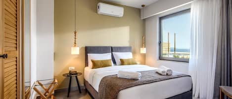 A modern hotel room with a large bed, a bedside table, a small side table, a wall-mounted air conditioner, and a window with a view of the sea.