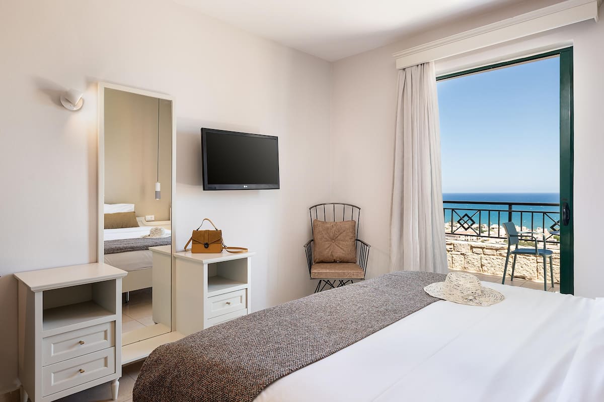 A hotel room with a bed, a TV, a chair, a desk, and a view of the beach.