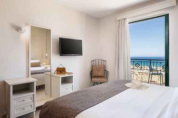 A hotel room with a bed, a TV, a chair, a desk, and a view of the beach.