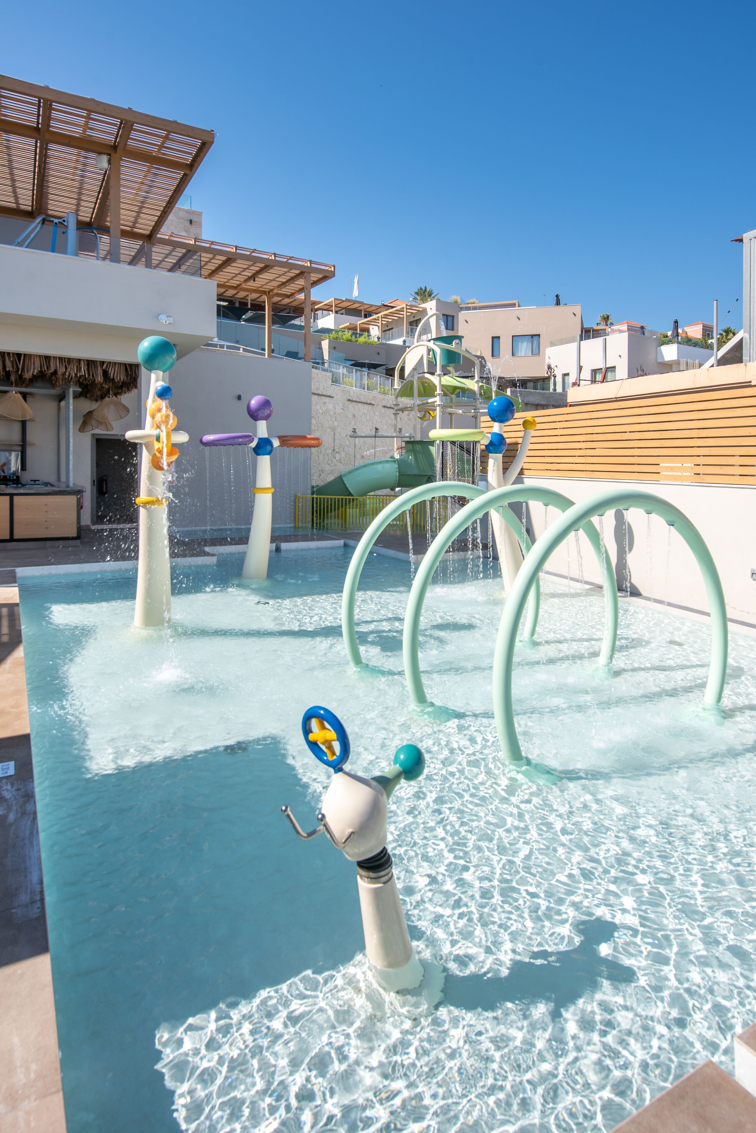 A water play area with multiple water jets and arches, surrounded by buildings and a clear sky.