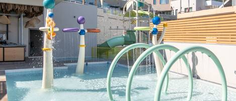 A water play area with multiple water jets and arches, surrounded by buildings and a clear sky.