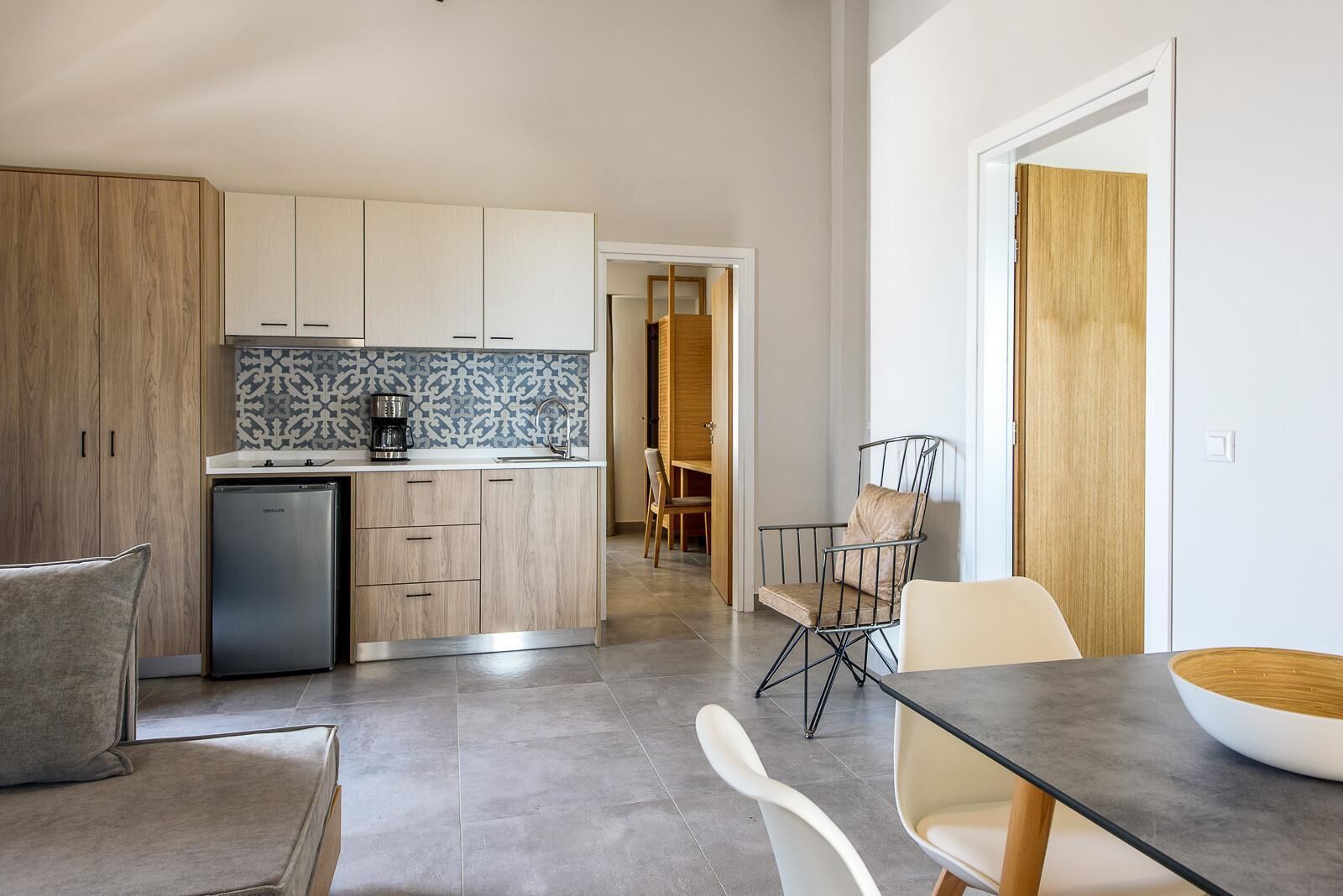 A modern kitchen with wooden cabinets, a built-in oven, and a coffee maker. There is a dining area with a table and chairs.