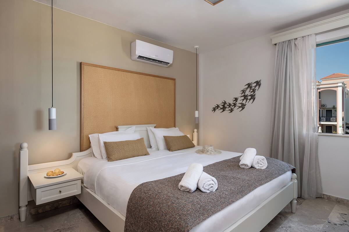 A neatly made bed with white linens and two rolled towels, a bedside table with a plate of cookies, a wall-mounted air conditioner, and a decorative wall piece.