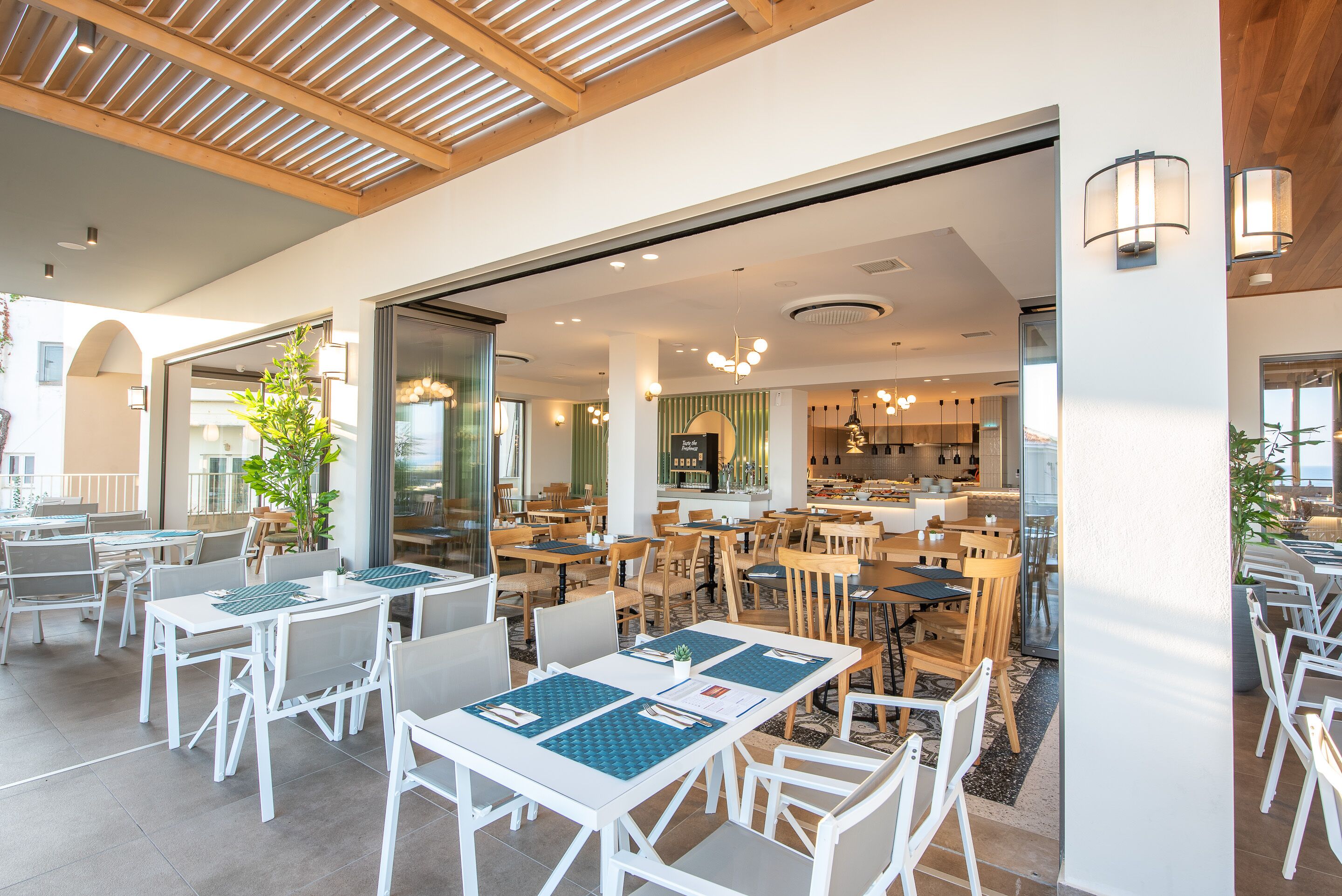 Un área de comedor al aire libre amplia, con mesas y sillas blancas, techo de madera y vista al área de la cocina al fondo.