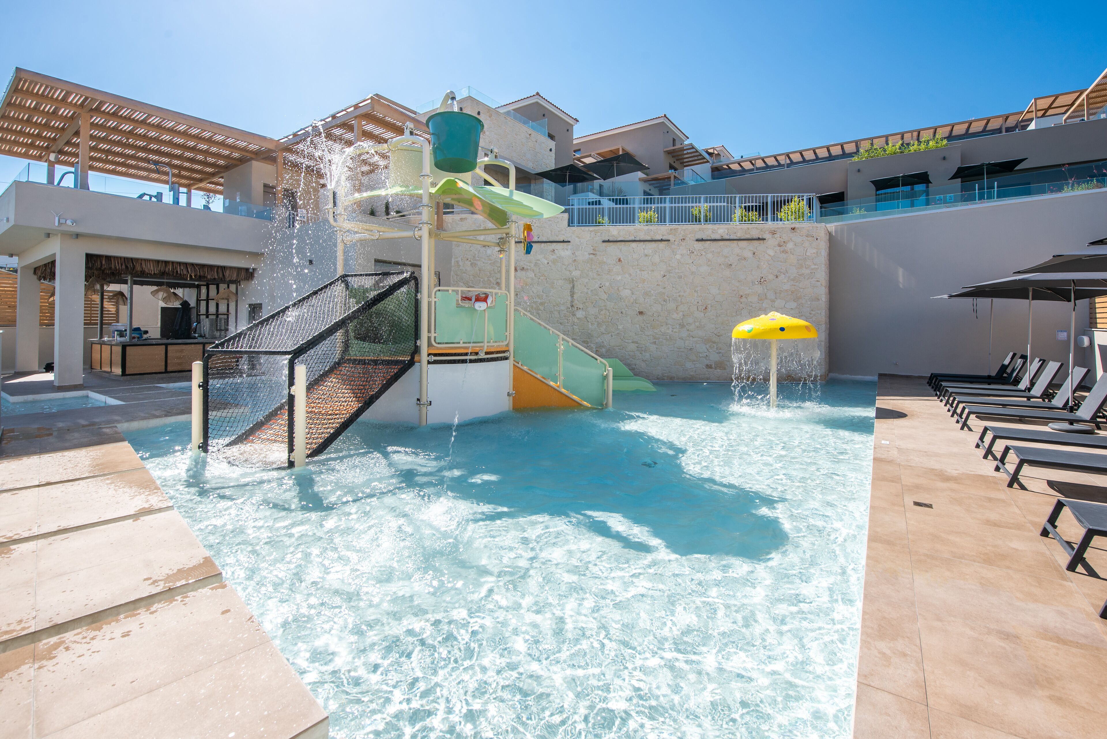 A water play area with slides and a pool, surrounded by lounge chairs and a building with balconies.