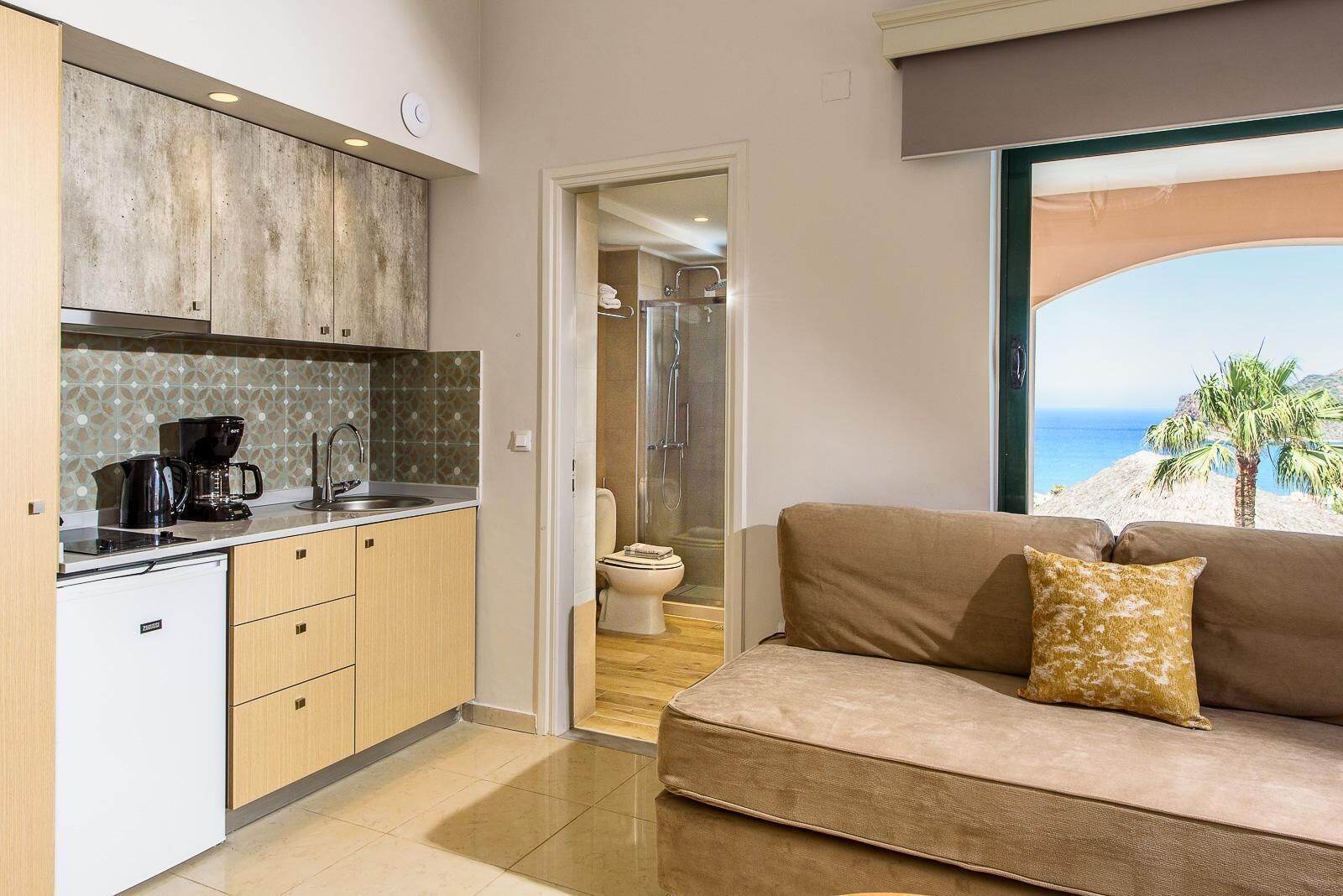 A modern kitchen with a coffee maker, a sink, and a toilet visible through an open door. A beige sofa with a patterned pillow is in the foreground, and a large window offers a view of the ocean.